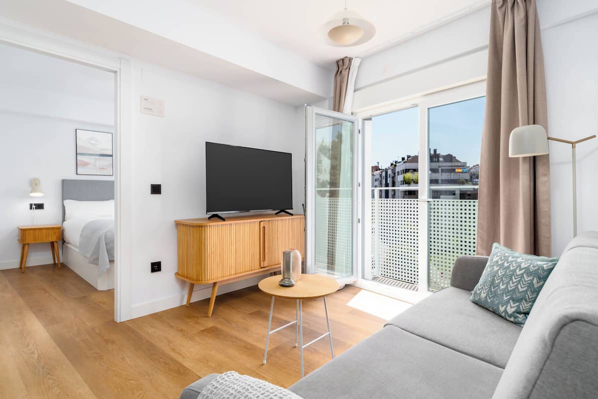 Un salon moderne avec un canapé, un téléviseur posé sur un meuble en bois, une petite table ronde et une porte-fenêtre coulissante donnant sur un balcon.
