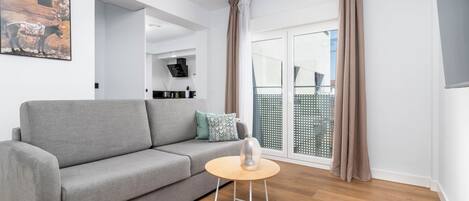 Ein modernes Wohnzimmer mit einem grauen Sofa, einem kleinen runden Tisch und einem großen Fenster mit Vorhängen.