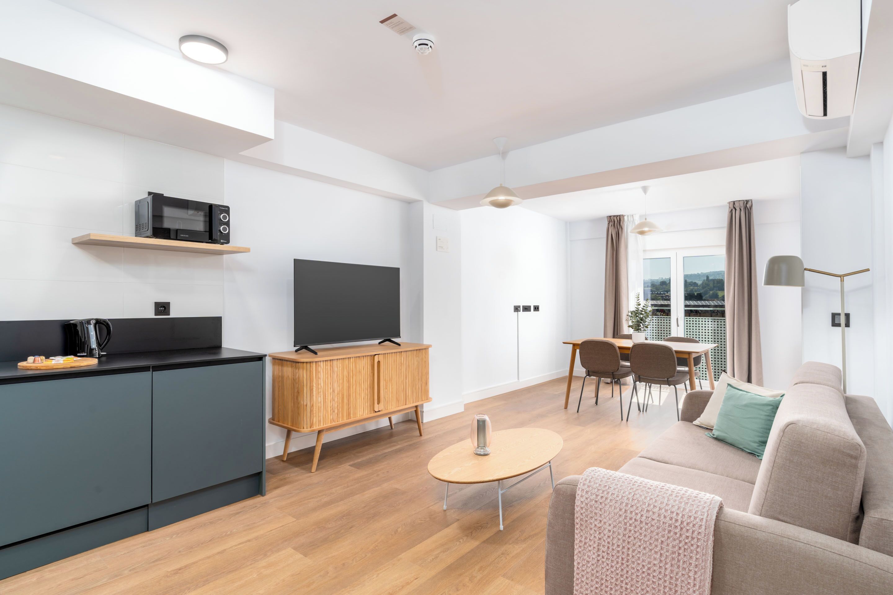 A modern living room with a sofa, coffee table, and a TV on a cabinet. There is a dining area with chairs and a table, and a balcony with a view.