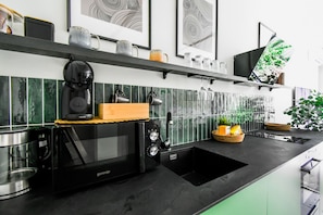 Una cucina moderna con un piano di lavoro nero, un forno a microonde integrato, una macchina da caffè e un lavello. Aggiunte decorative sullo scaffale sopra il microonde, tra cui una tazza, una pianta e una cornice con foto.