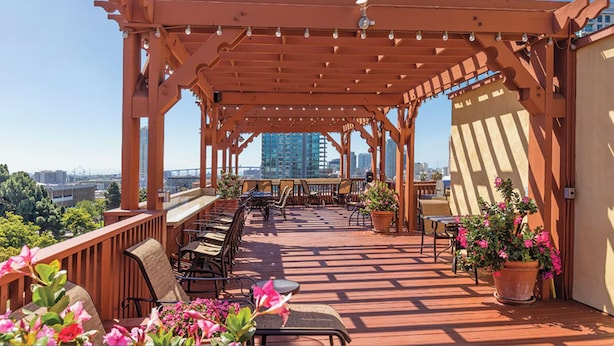 A covered outdoor seating area with wooden chairs and tables, potted plants, and a view of the cityscape. - Explore San Diego right outside your door. (San Diego)