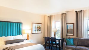 A hotel room with a bed, a sofa, a desk, and two chairs. There are two windows with curtains, and a painting on the wall. - Explore San Diego right outside your door. (San Diego)