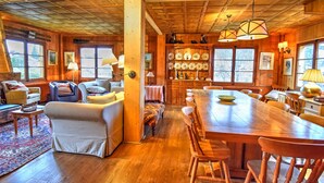 Acogedor interior de cabaña de madera con comedor, sala de estar con sofá y sillones, y cocina con armarios de madera y exhibición de platos.