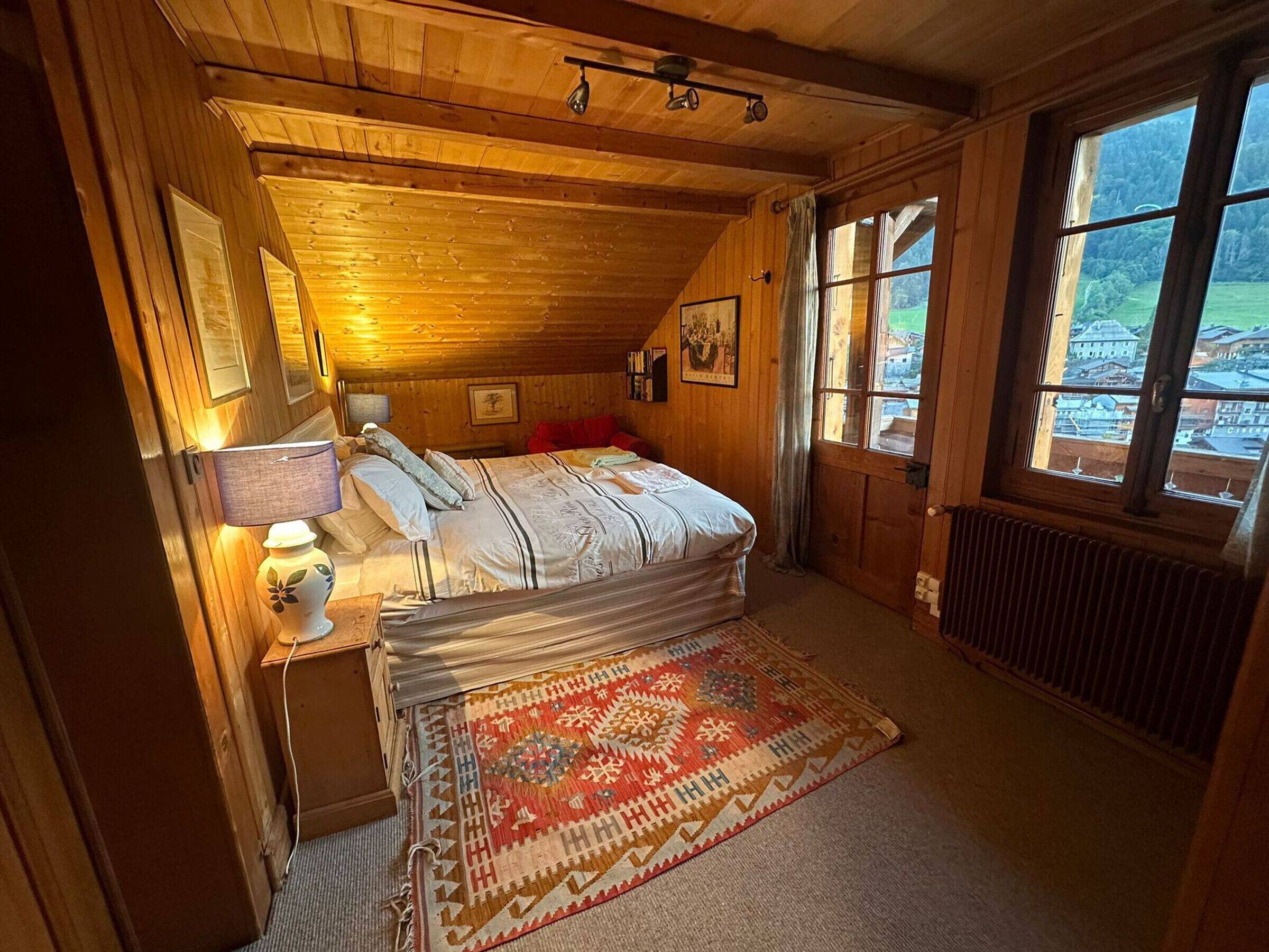 A cozy bedroom with a wooden ceiling, a large window, a bed with a patterned bedspread, a bedside table with a lamp, and a decorative rug.