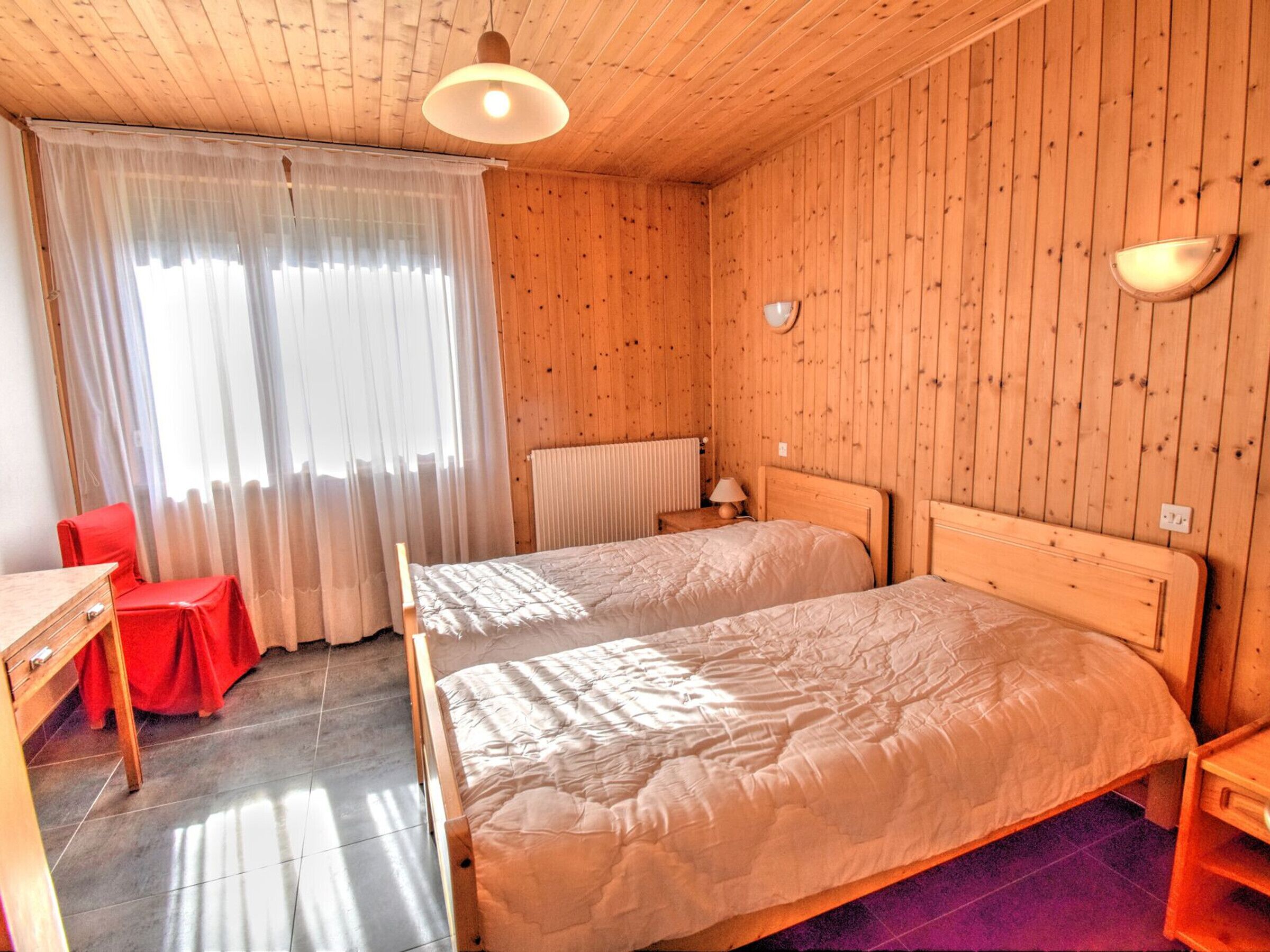 A wooden-paneled room with two beds, a desk, a chair, and a window with curtains.