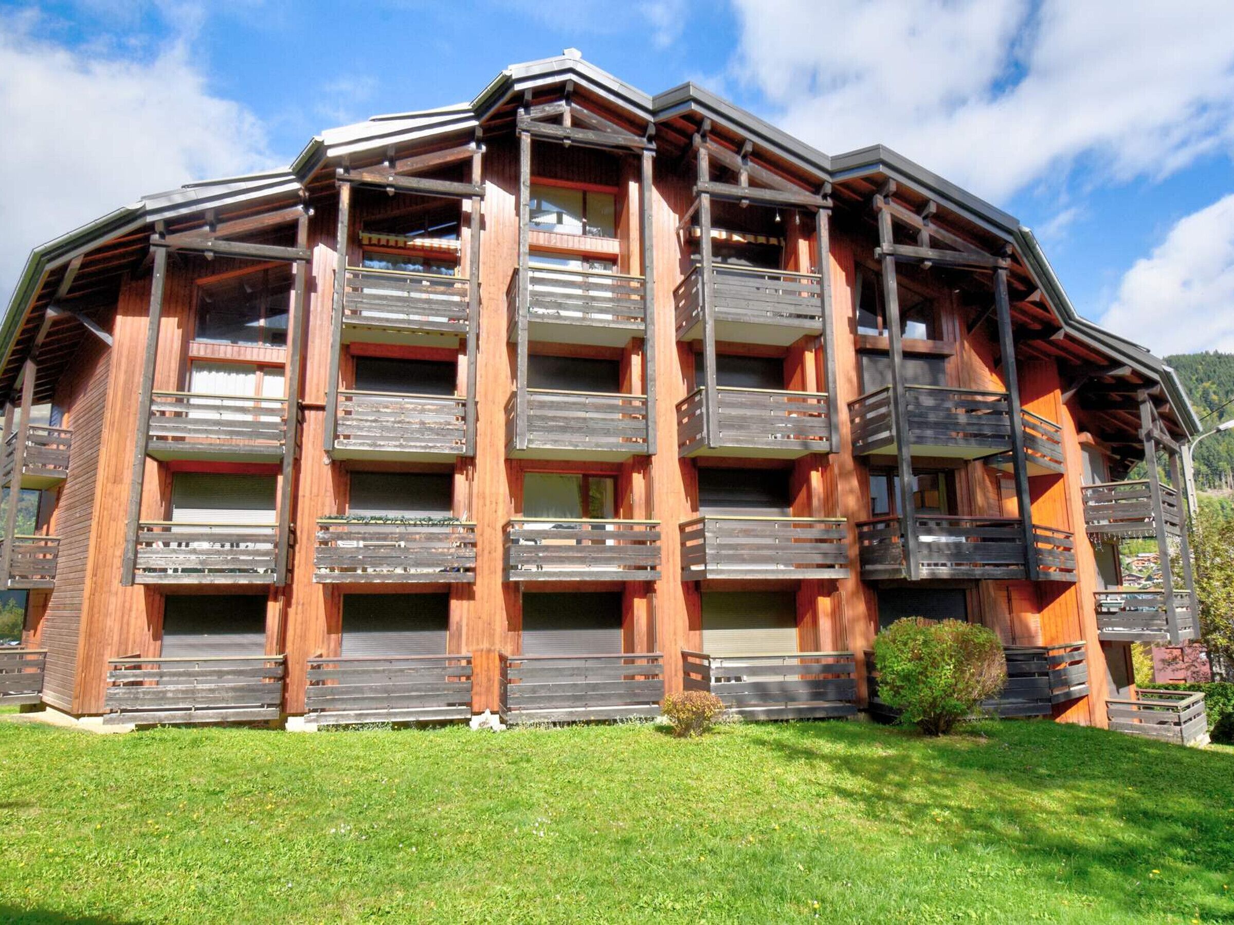 A multi-story wooden building with balconies, likely a hotel or guesthouse, surrounded by a grassy area and a clear sky.