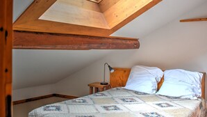 Et soveværelse med skråvæg, træbjælker, en seng med hvide sengetøj og et lille natbord med en lampe.