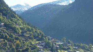 Een bergachtig dorp, met huizen verscholen tussen de bomen, een heldere lucht en besneeuwde bergtoppen op de achtergrond.