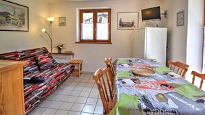 En hyggelig stue med sofa, spisebord og stole. Der er et vindue med gardiner, en lampe og et fjernsyn på væggen.