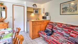 En kompakt opholdsstue med et tekøkken, en sofa med et mønstret betræk, et træspisebord med stole og et spejl.