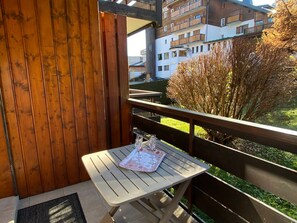 A balcony with a wooden railing, a small table with a glass of water, and a view of a building with balconies.