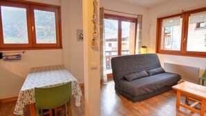 Ein Zimmer mit Holzboden, einem grünen Sessel, einem Tisch mit gemustertem Tischläufer, einem grauen Sofa und einer Holzbank.