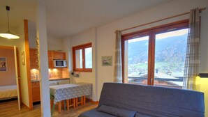 Ein gemütliches Wohnzimmer mit grauem Sofa, Holzboden, einer Essecke mit Tisch und Stühlen, einer Küche mit Mikrowelle und einem großen Fenster mit Blick auf eine gebirgige Landschaft.