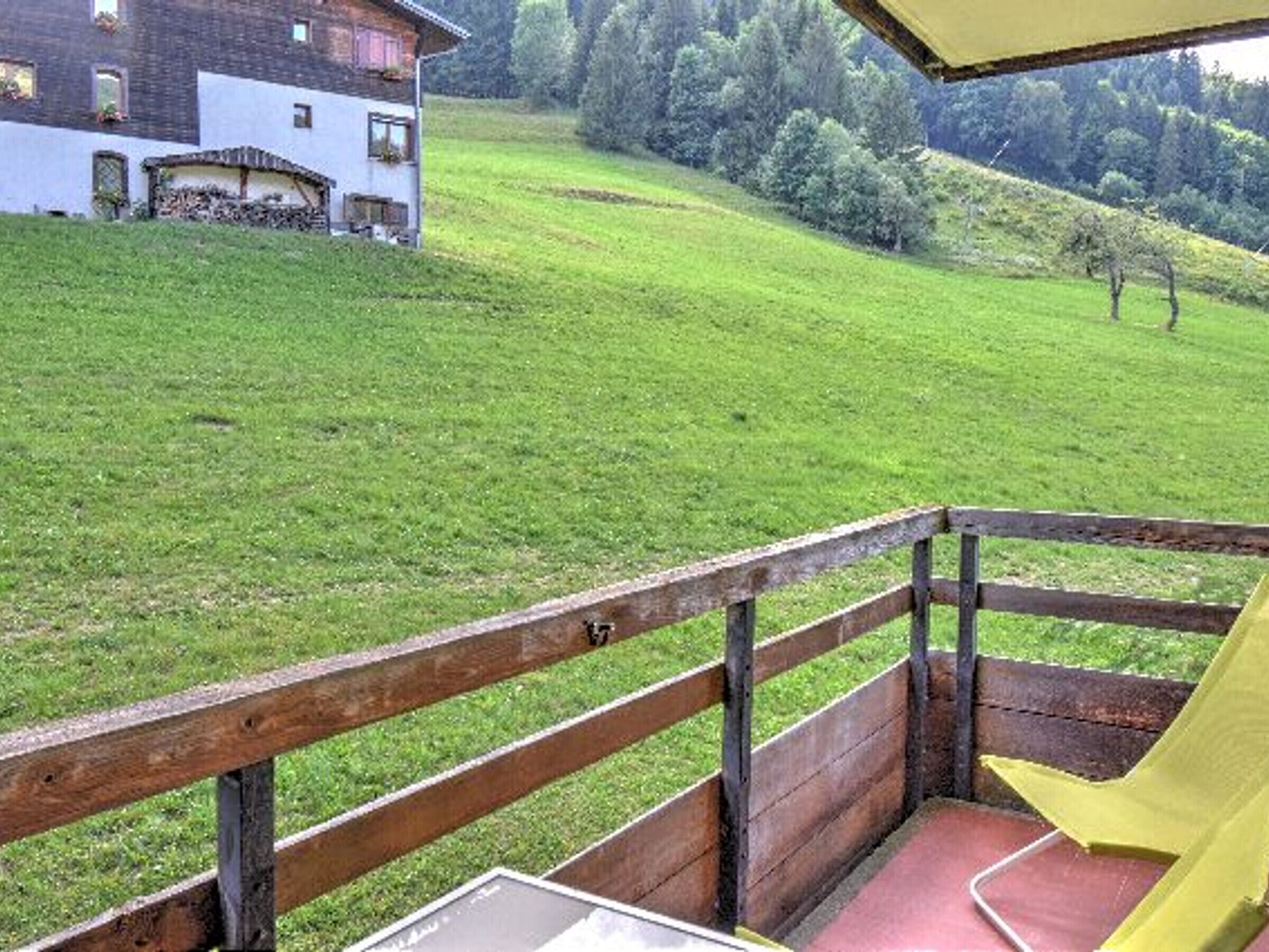 Eine Holzterrasse mit gelbem Markisenstoff, einer roten Bank und Blick auf ein Gebäude und eine grüne Landschaft.