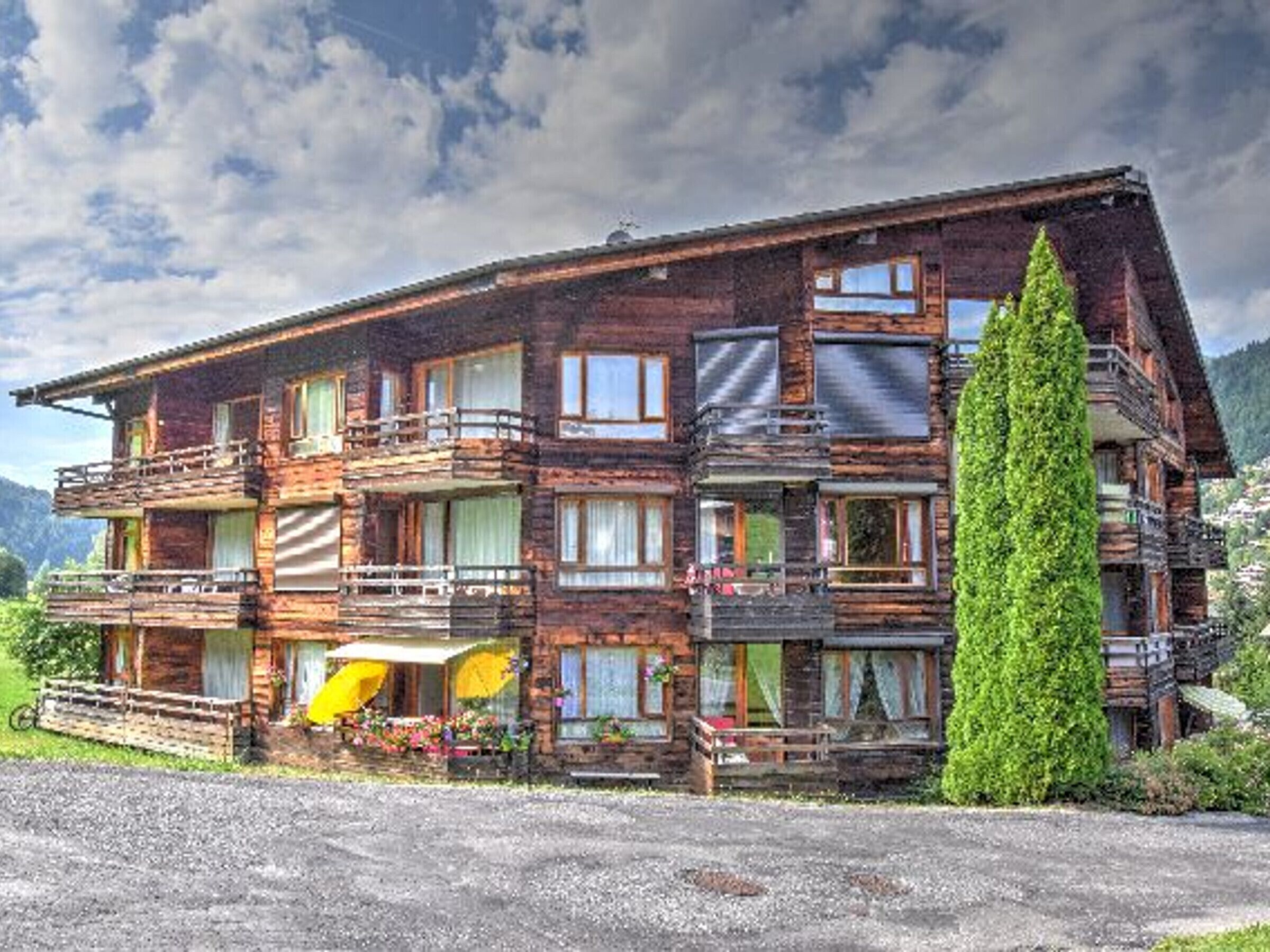 A multi-story wooden chalet with balconies, a gravel driveway, and a large coniferous tree in the foreground.