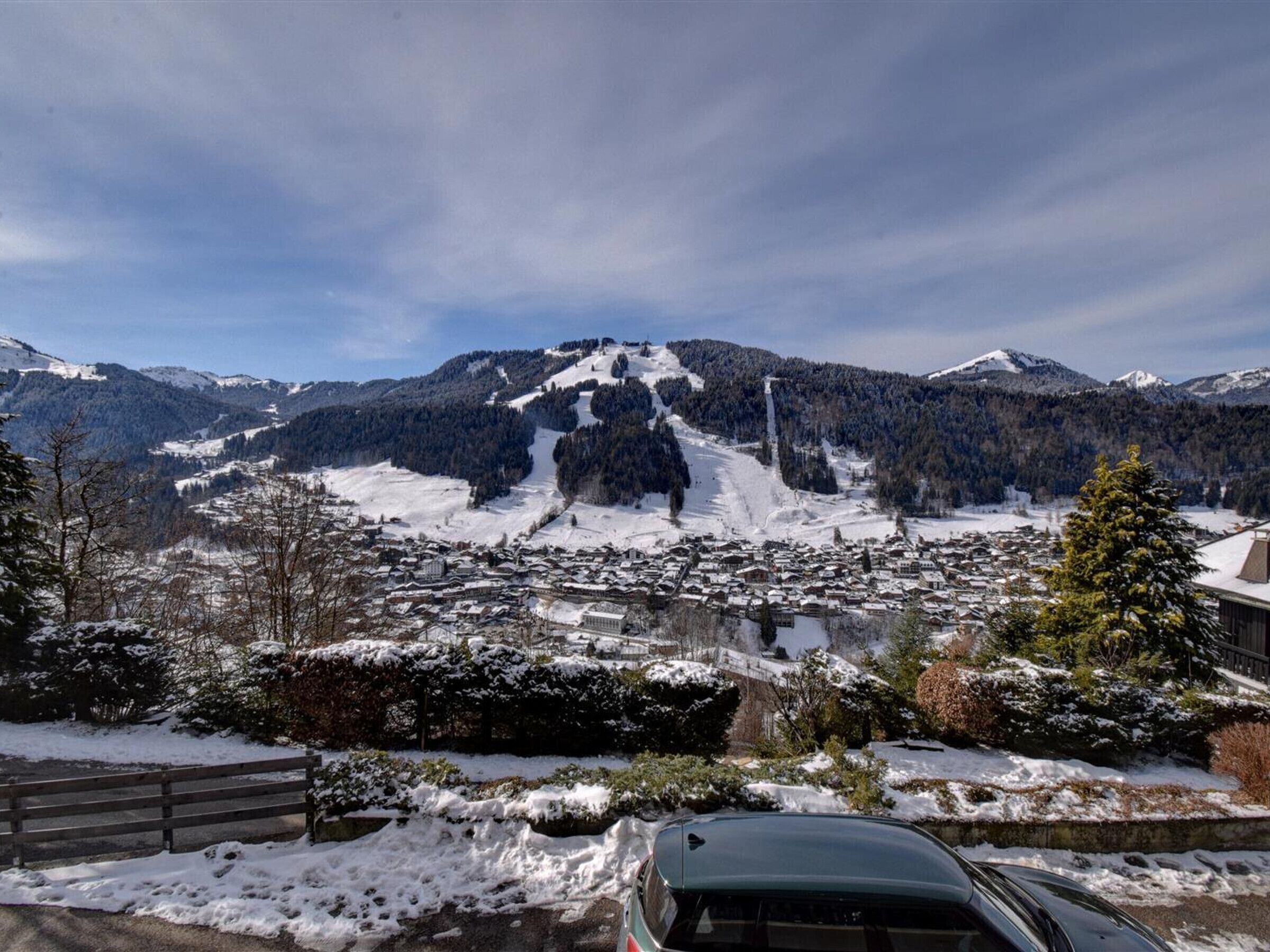 A snow-covered mountain town with a ski slope, a car, and a wooden fence.