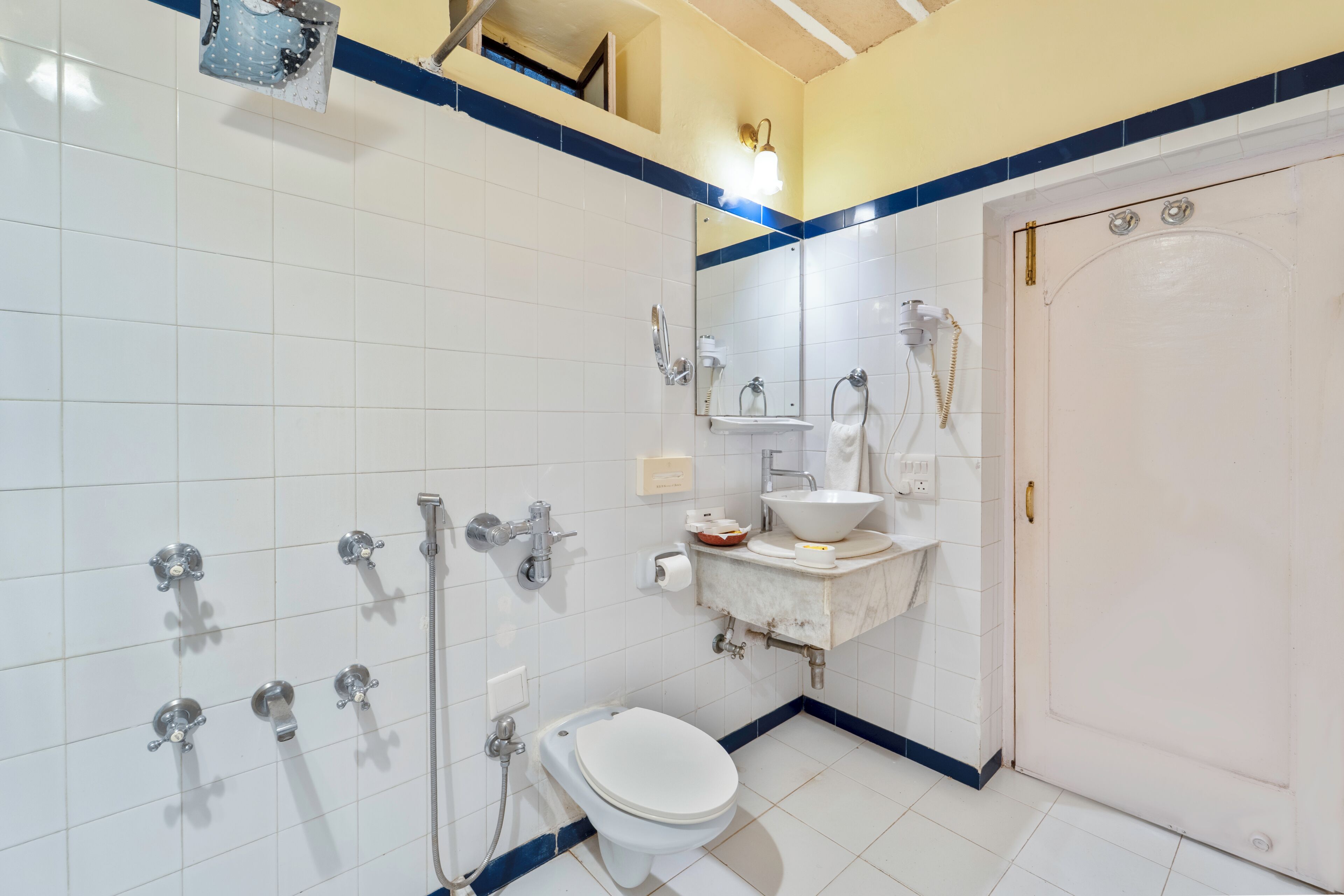A bathroom with white tiled walls, a white toilet, a sink with a mirror above it, and a shower with multiple faucets.