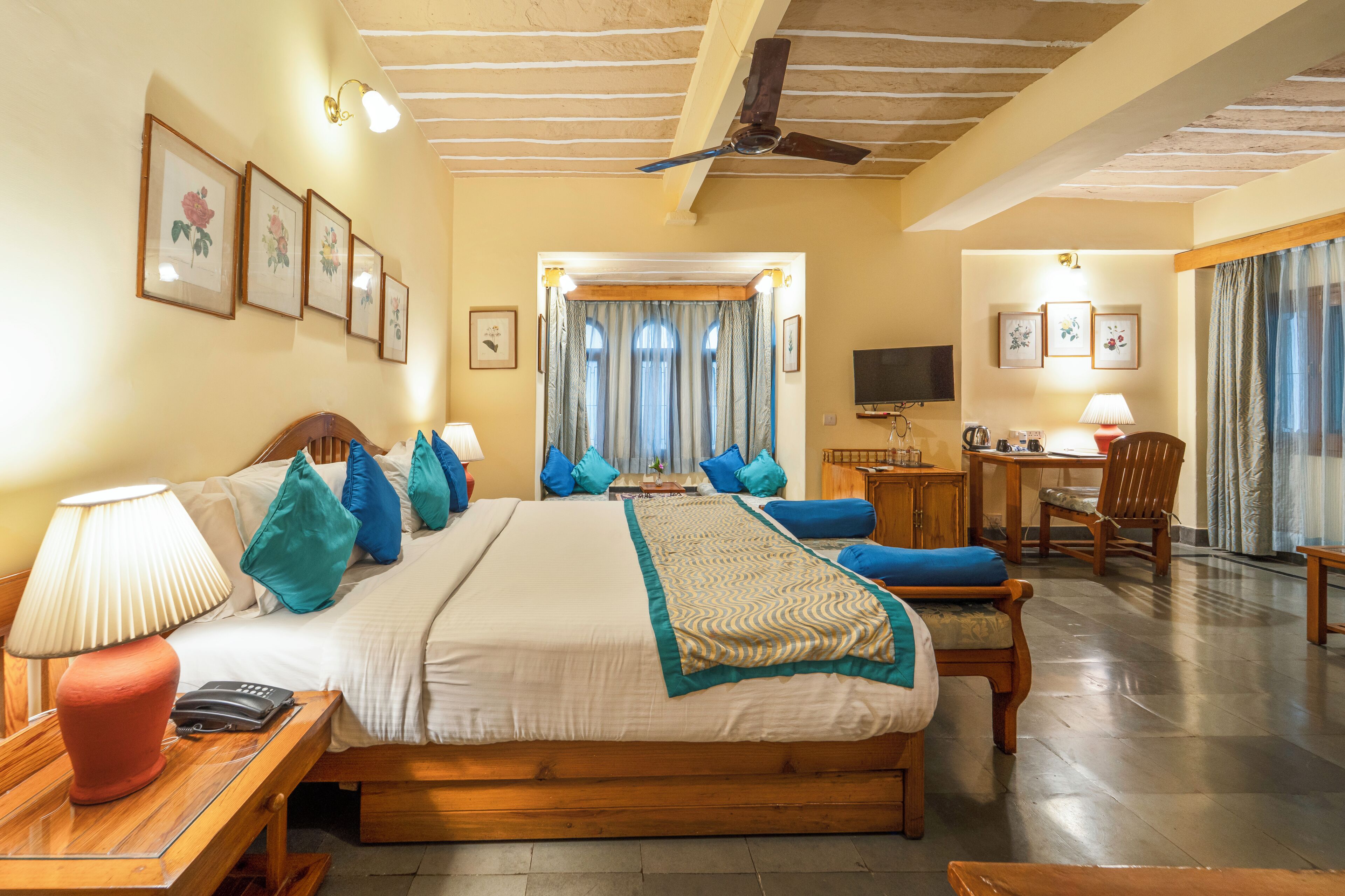 A hotel room with a bed, bedside tables, a desk, a chair, a television, and a ceiling fan.