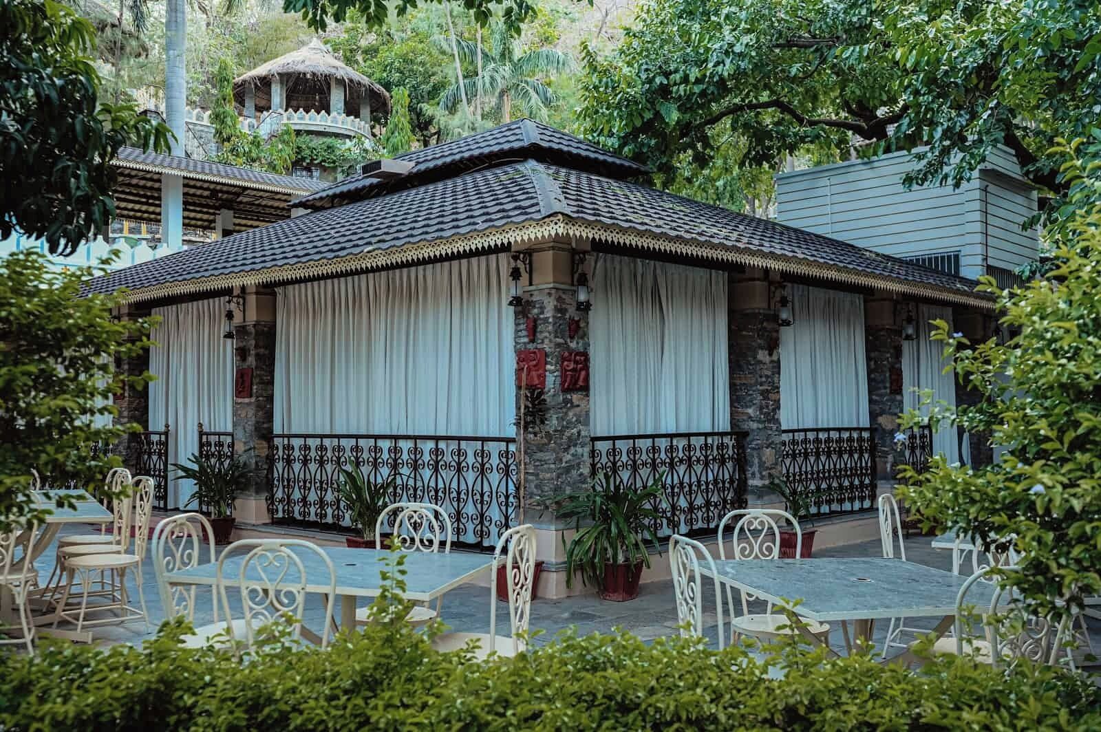 A traditional building with a tiled roof and white curtains, surrounded by potted plants and outdoor seating.