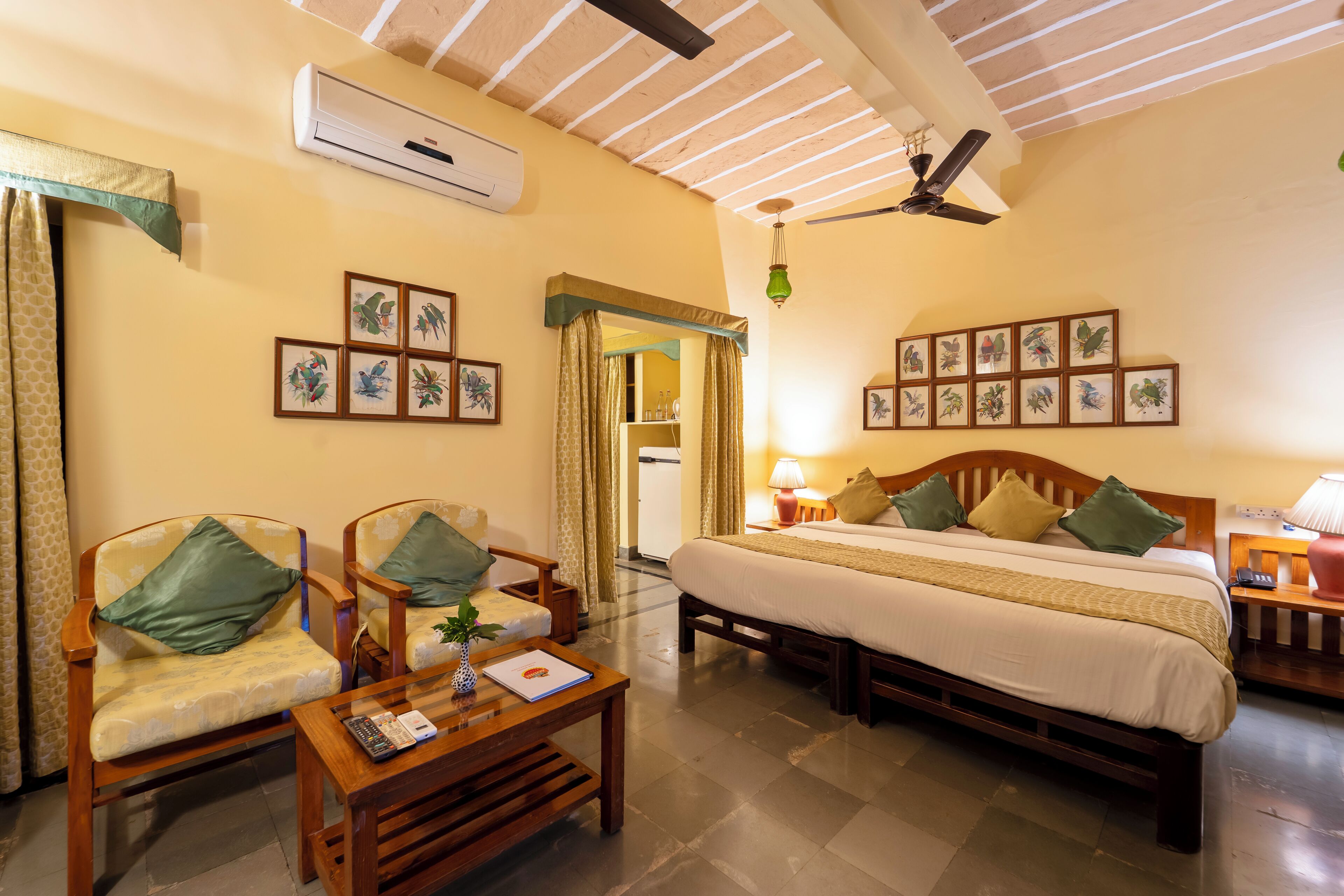 A hotel room with a bed, two chairs, a table, an air conditioning unit, a ceiling fan, and framed bird pictures on the wall.