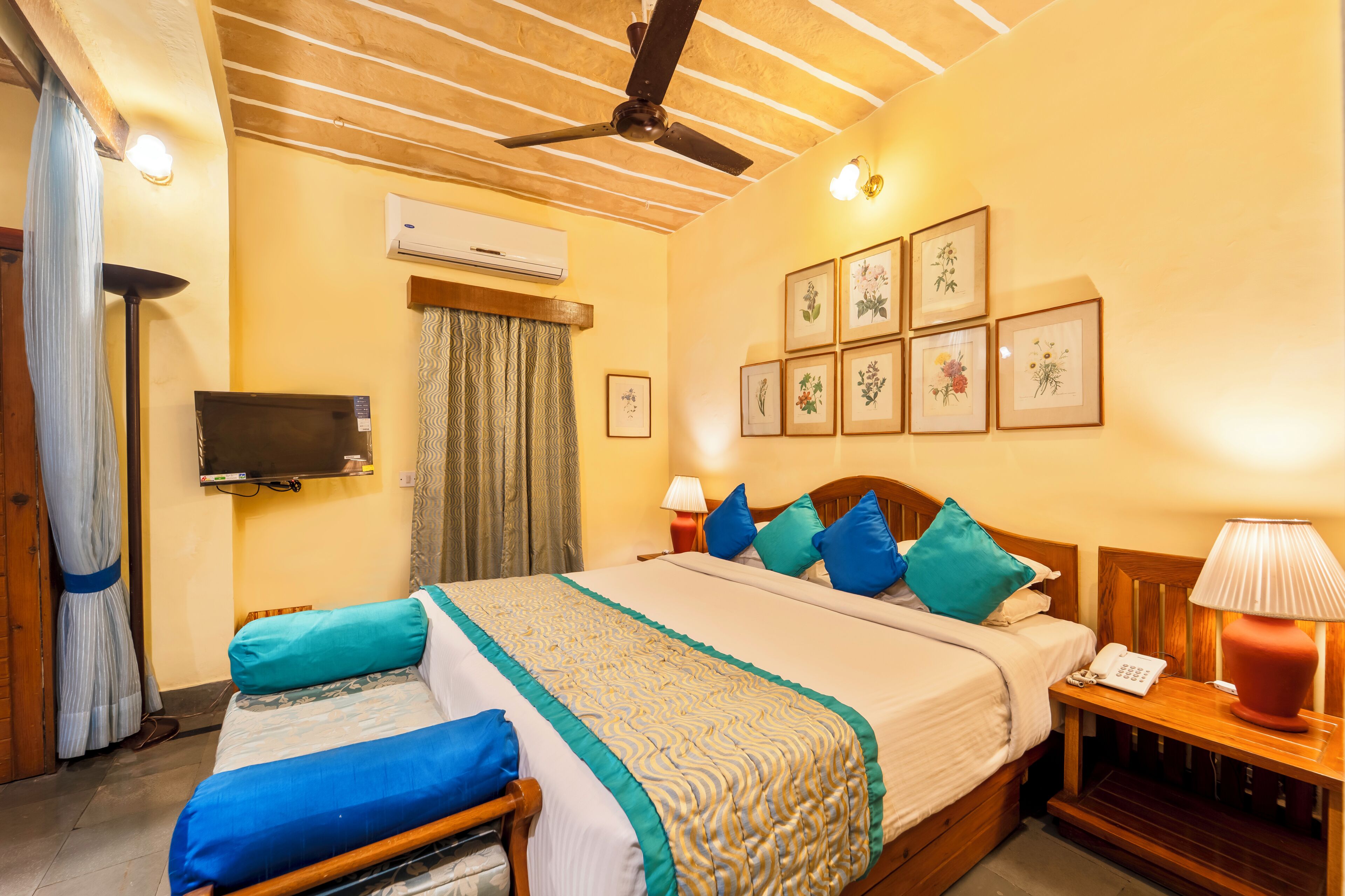 A hotel room with a bed, a ceiling fan, an air conditioning unit, a television, a window with curtains, and framed botanical prints on the wall.