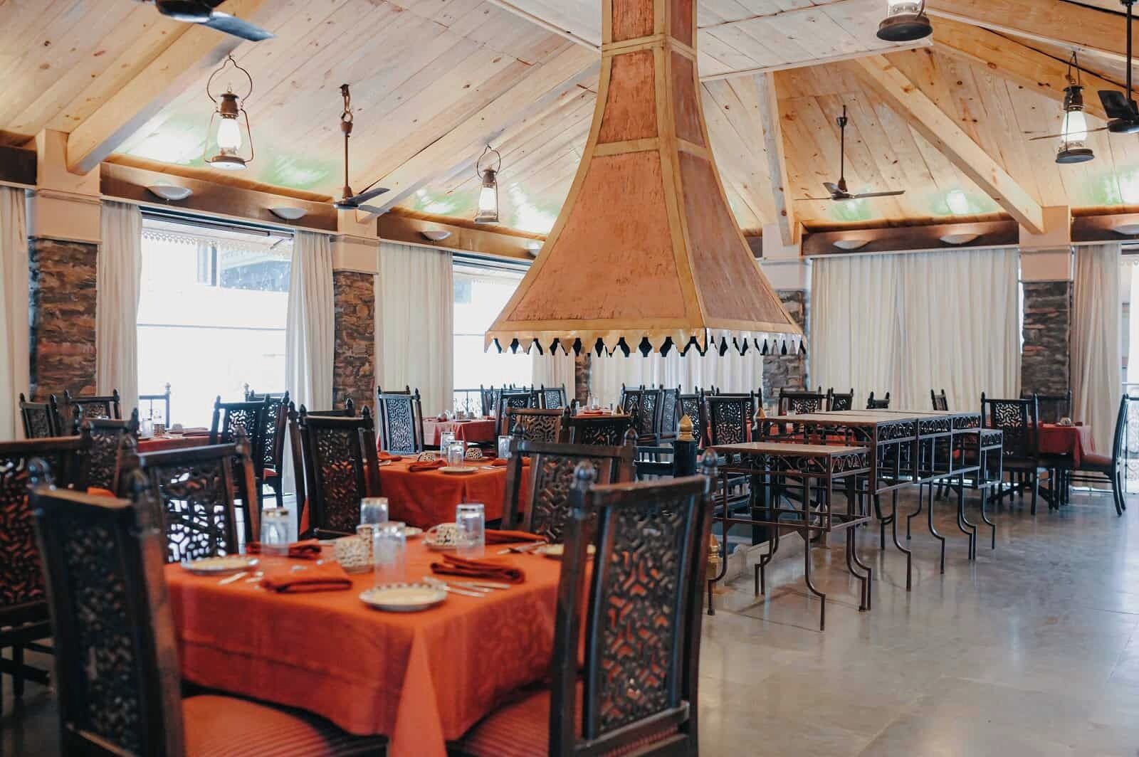 A spacious dining area with high wooden ceilings and large windows, featuring tables set with orange tablecloths and ornate chairs.