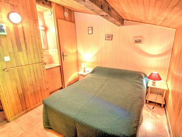 A small, cozy room with a bed, a wooden door, a small table with a lamp, and a wall-mounted mirror.