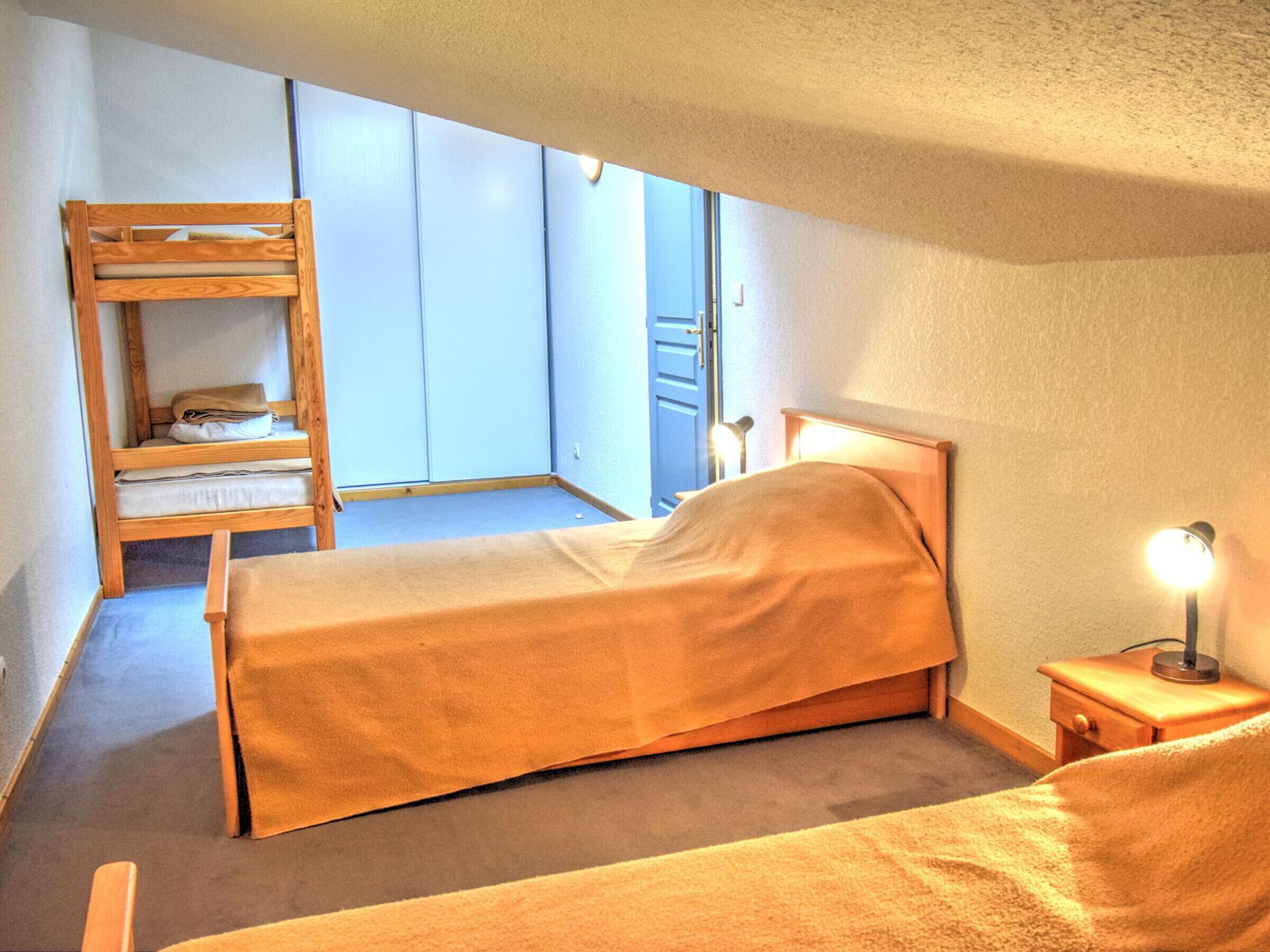 Een knus kamertje met twee bedden, inclusief een stapelbed van hout, een nachtkastje met lamp, en een raam met uitzicht.
