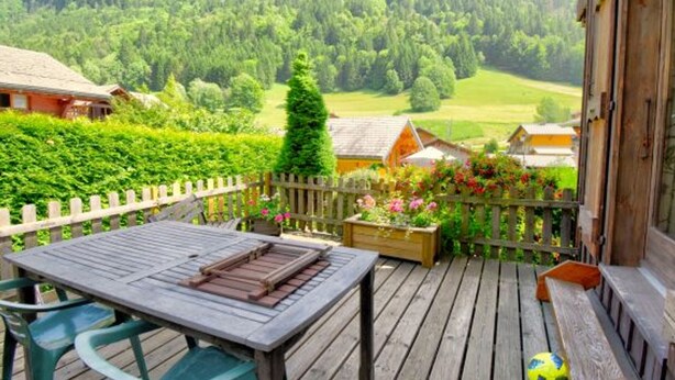 Eine Holzterrasse mit Tisch und Stühlen, umgeben von einem Holzzaun, mit Blick auf eine üppige grüne Landschaft mit Häusern und Bergen im Hintergrund.