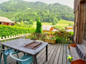 A wooden deck with a table and chairs, surrounded by a wooden fence, overlooking a lush green landscape with houses and mountains in the background.