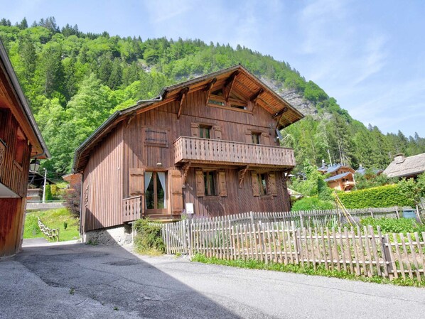A wooden chalet with a balcony, set in a mountainous area with a wooden fence and greenery.