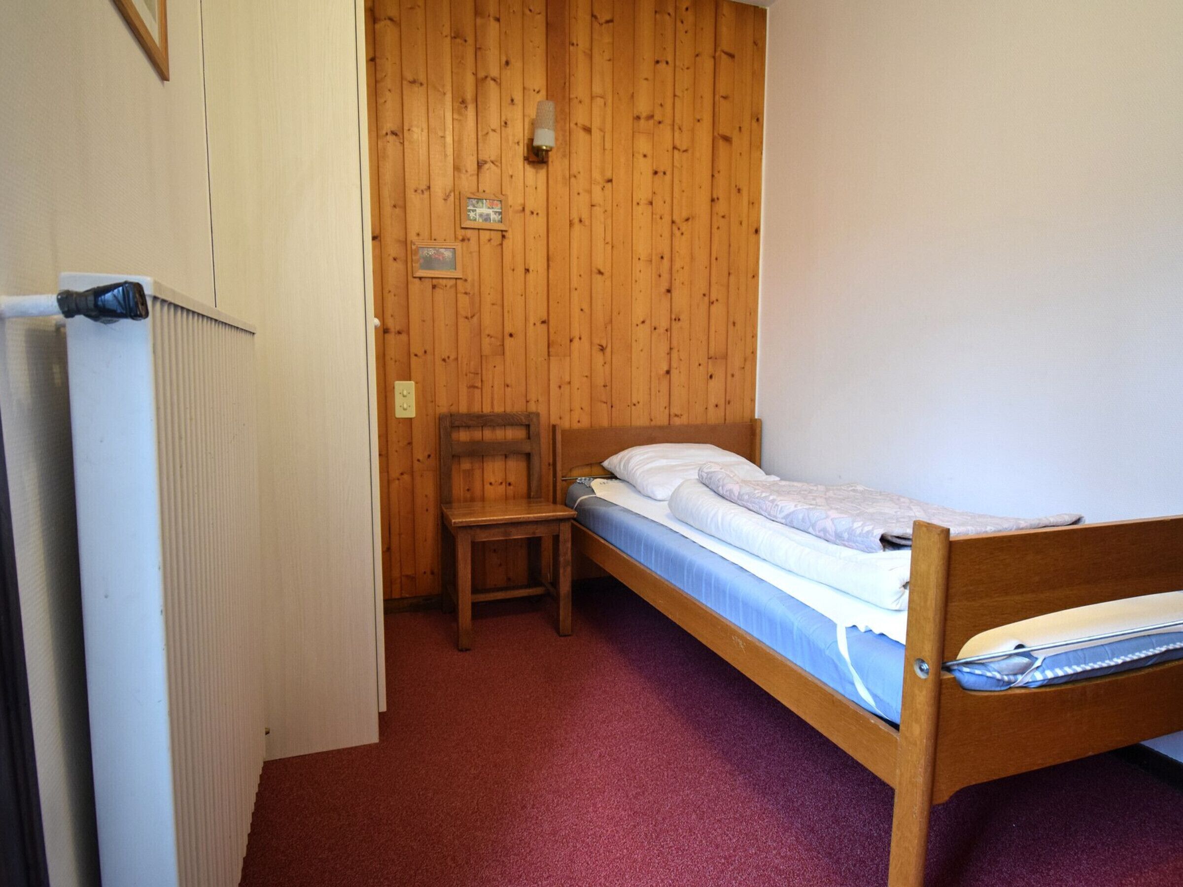 Une petite chambre avec un lit simple, des murs en bois, une chaise en bois et un radiateur.