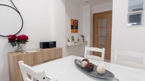 Ein Essbereich mit weißem Tisch, Stühlen, einer Zierplatte, einer Blumenvase und einem Schrank mit Lautsprecher.