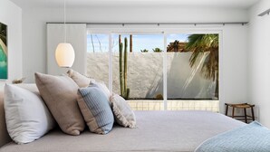 A bedroom with a large bed, multiple pillows, a hanging light fixture, and a view of a beach through large windows.