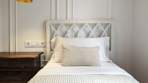 Een net opgemaakt bed met witte beddengoed en een gestructureerd kussen, een nachtkastje van hout met lades, en een hanglamp.