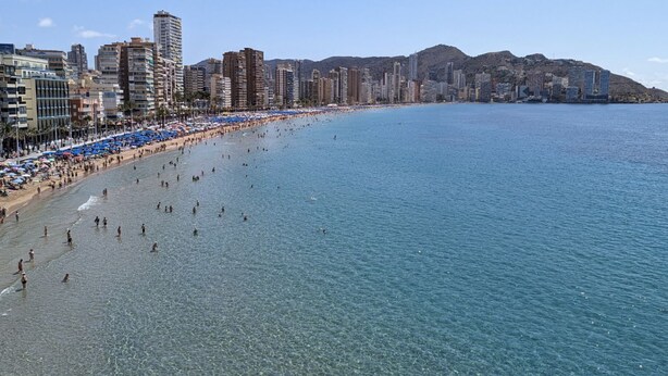 Een strand met helderblauw water, een zandstrand en een stadsbeeld met hoge gebouwen op de achtergrond.