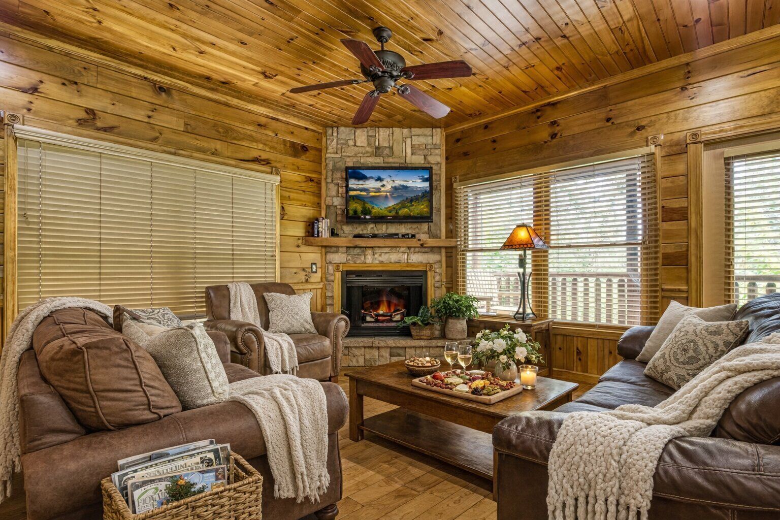 A cozy cabin interior with a stone fireplace, leather couches, a television, and a ceiling fan.