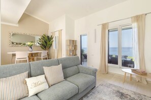 Una sala de estar con un sofá gris, una mesa de comedor y vista al mar a través de grandes ventanales.
