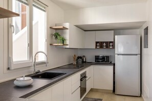 A modern kitchen with white cabinets, a black countertop, a stainless steel refrigerator, and a window with blinds. - Seaside Home where Villasimius's sea becomes home (VILLASIMIUS)