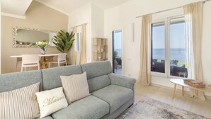 Una sala de estar con un sofá gris, una mesa de comedor y vista al mar a través de grandes ventanales.