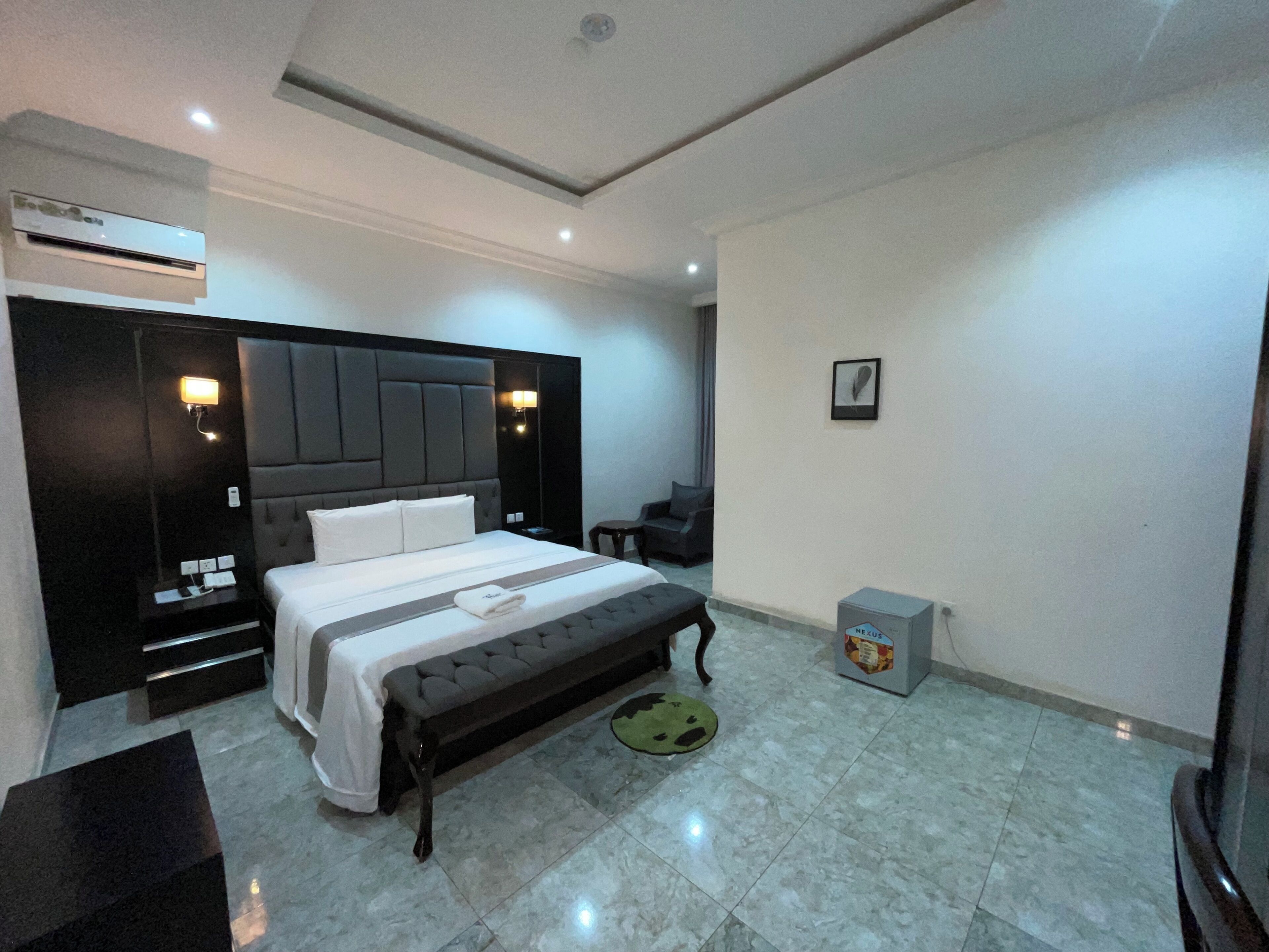 A modern hotel room with a large bed, a bedside table, a nightstand, an air conditioning unit, a small desk, a chair, and a trash bin.