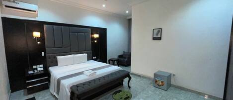 A modern hotel room with a large bed, a bedside table, a nightstand, an air conditioning unit, a small desk, a chair, and a trash bin.