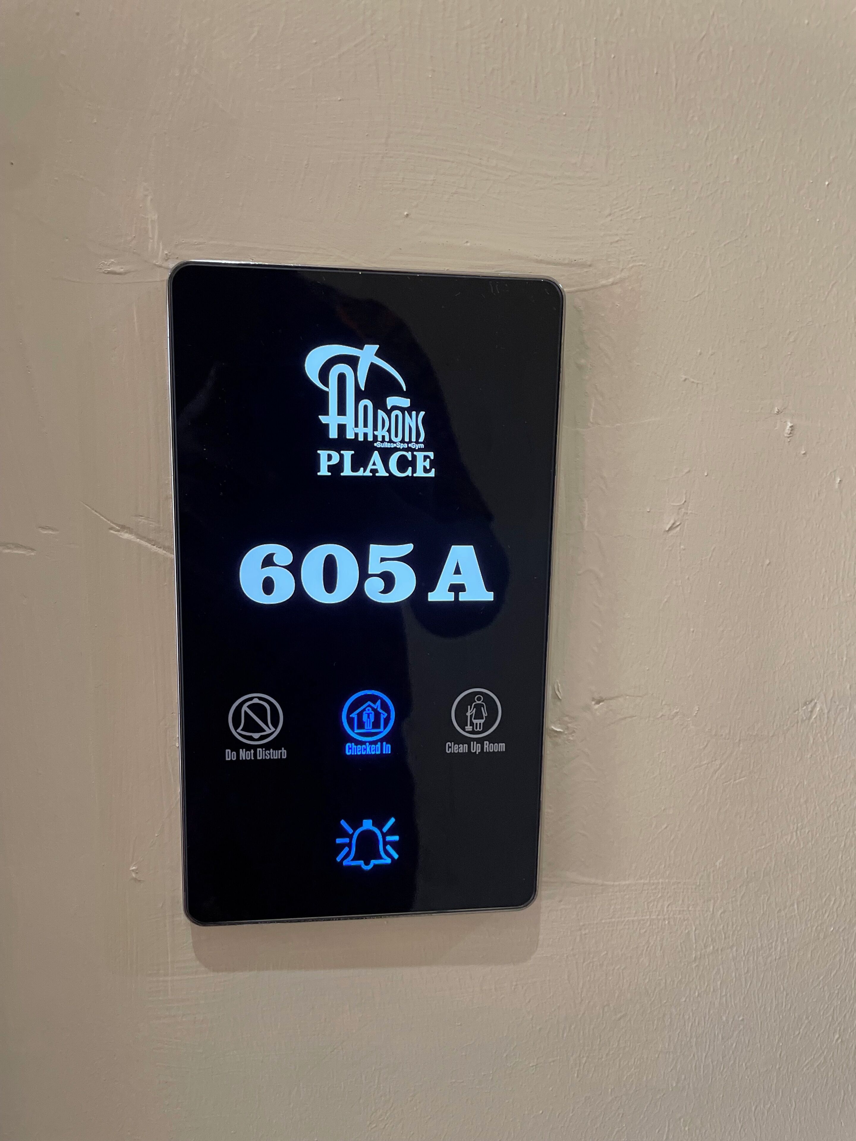 Pantalla digital que muestra el texto "HARRONS PLACE" y "605A", indicando un número de habitación. Incluye iconos de "No molestar", "Registrado/a" y "Limpiar habitación". 