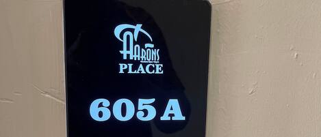 Pantalla digital que muestra el texto "HARRONS PLACE" y "605A", indicando un número de habitación. Incluye iconos de "No molestar", "Registrado/a" y "Limpiar habitación".