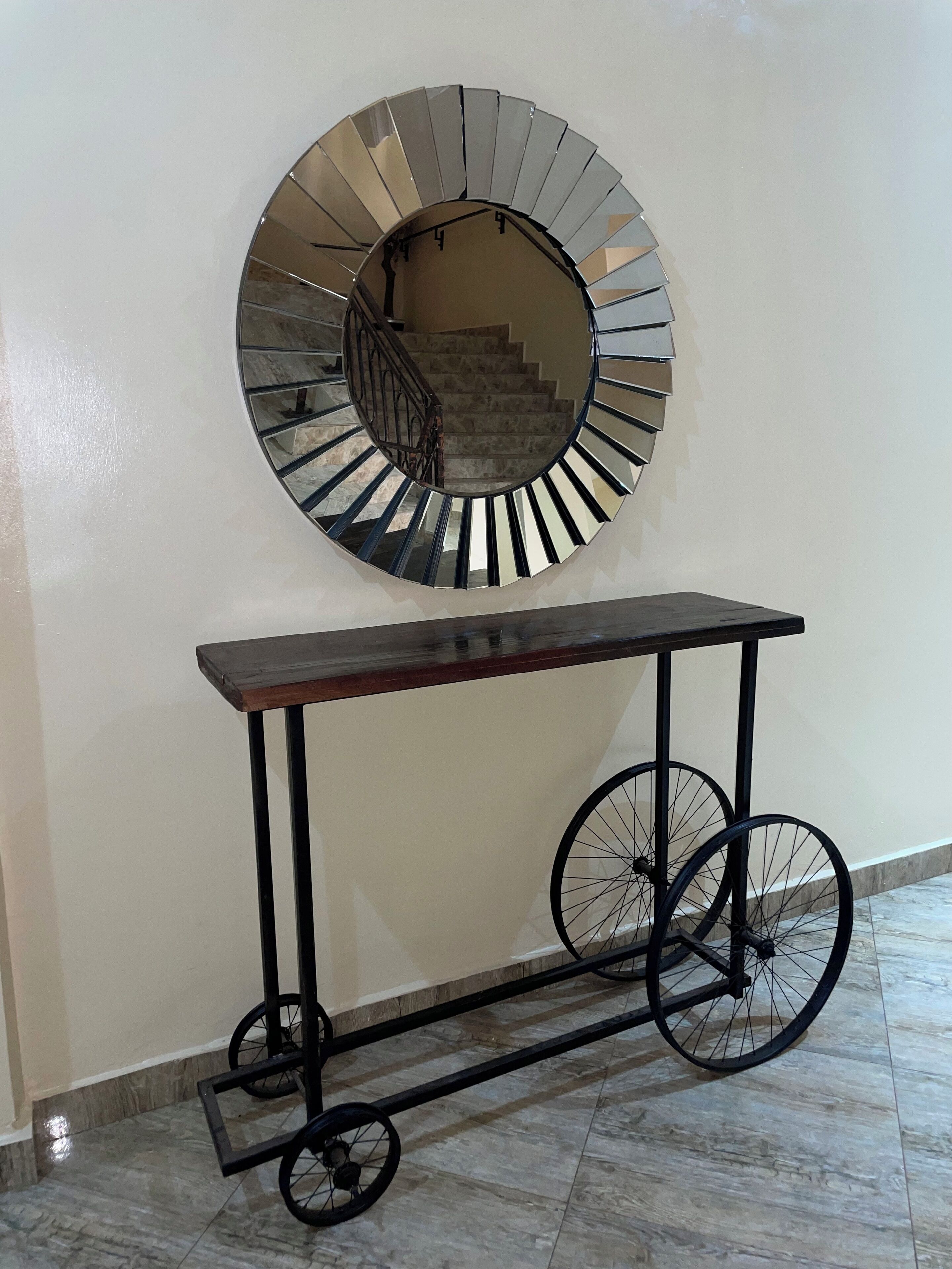A round mirror with a metallic frame, a wooden console table with wheels, and a bicycle wheel attached to the table.