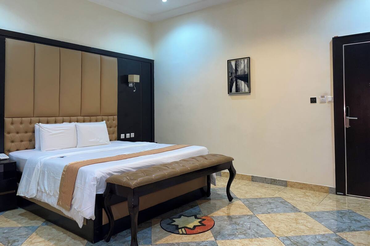 Une chambre d’hôtel avec un grand lit, un banc, une table de chevet, une lampe fixée au mur, une porte et un tableau encadré accroché au mur.