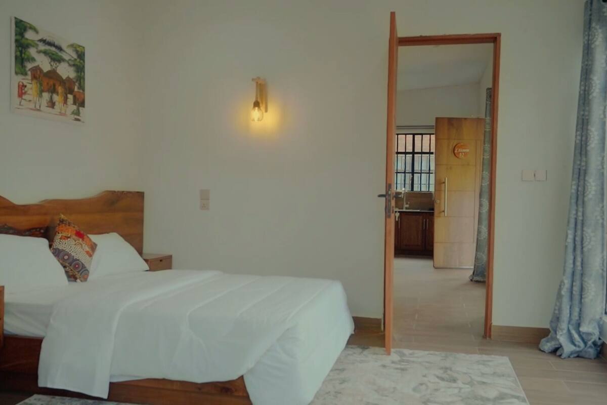 Un dormitorio con cama, mesita de noche, lámpara de pared y puerta que da a una cocina.