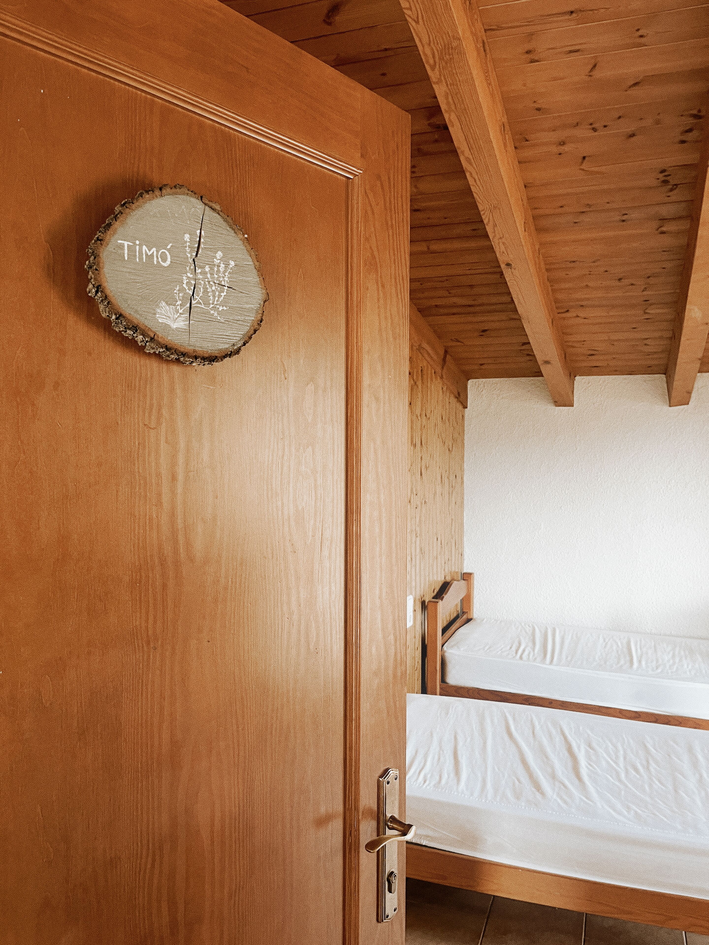 A wooden door with a circular wooden sign that reads "Timo" and a crack. There is a wooden bed frame with white bedding.