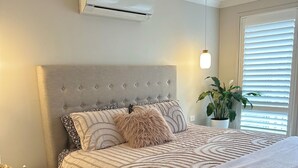 Une chambre à coucher avec une tête de lit capitonnée, un lit avec une literie à motifs, une table de chevet avec une lampe et un appareil de climatisation placé au-dessus du lit.