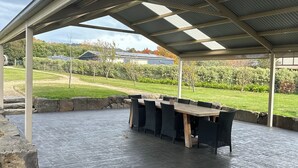 Un patio couvert doté d’une longue table et de chaises en bois, entouré d’une bordure en pierre et d’un sol pavé.
